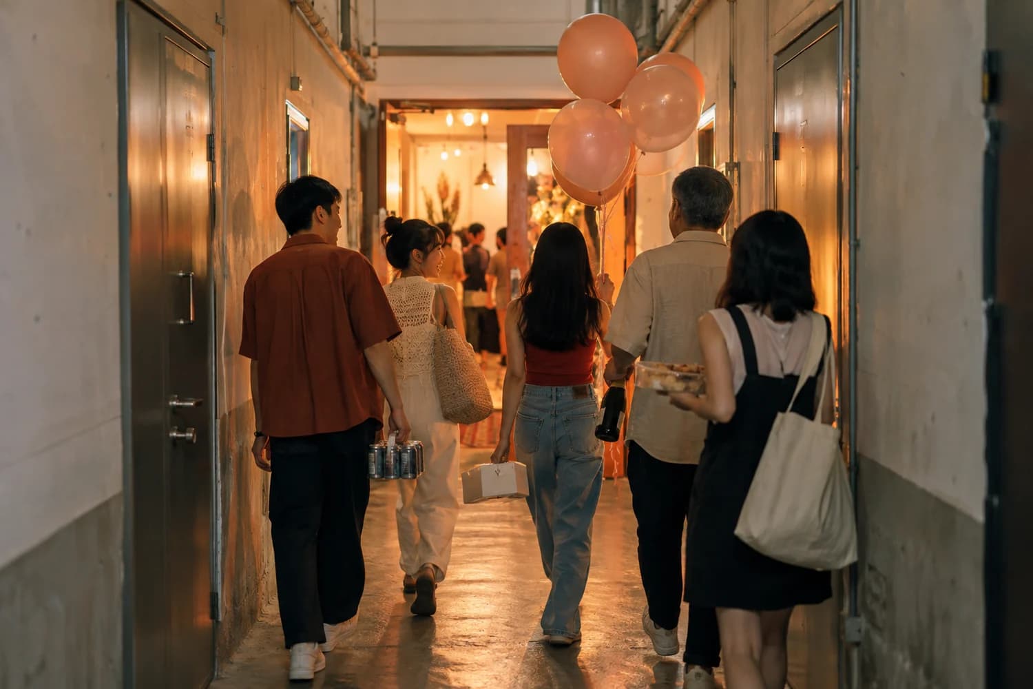 Friends walking down a long industrial building corridor towards a unit door with drinks and balloons
